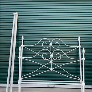 Può includere: Struttura letto in metallo bianco con volute ornate e una testata con un design decorativo. Il telaio è posizionato davanti a un portone verde del garage.