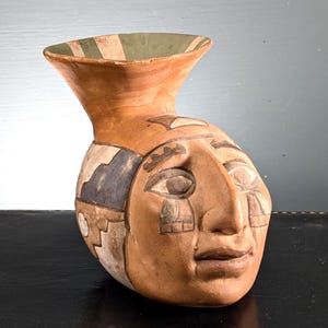 May include: A hand-painted ceramic vase shaped like a human face. The vase is primarily tan, with painted details in brown, blue, and white. The rim is a lighter tan with green and white stripes. The face has detailed eyes and a calm expression, with a serene look.