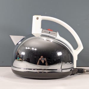 May include: A vintage General Electric electric kettle with a shiny chrome finish. It has a white handle and spout, a black base, and a red button. The kettle is designed with a classic, retro aesthetic.