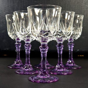 May include: A set of six crystal wine glasses, each with a clear bowl and a vibrant purple stem and base. The glasses feature a detailed diamond-cut pattern. The stems have an ornate design, and the bases are circular. The glasses are grouped together.