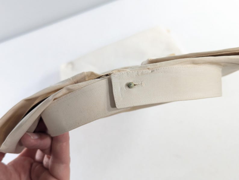 May include: Close-up of a beige dress shirt collar with a buttoned closure. The collar is folded over, revealing the inside lining. The shirt appears to be made of a light, possibly cotton, fabric.