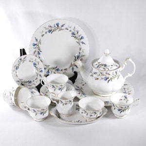 May include: A white porcelain tea set with a floral pattern of blue and purple berries and green leaves. The set includes a teapot, cups, saucers, plates, and a creamer, all with gold edging.