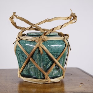 May include: A teal-colored ceramic jar with a woven bamboo handle and lattice design. The jar has a textured surface and a light-colored base. The bamboo handle is twisted and secured to the jar with twine.