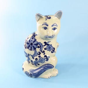 以下が含まれることがあります： クリーム色のベースに青い花柄が施された陶器製の猫の置物。猫は座った姿勢で、尻尾を体の横に巻き付けています。青く描かれた目、ひげ、眉毛が細部まで表現されています。