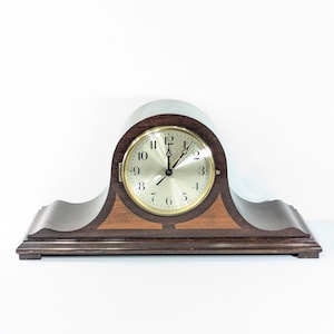 Peut inclure: Une horloge de manteau en bois brun foncé avec un cadran doré et un dessus incurvé. L'horloge a une base rectangulaire avec un motif ondulé décoratif. Le cadran affiche les chiffres de 1 à 12.