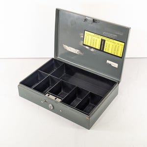 Puede incluir: Una caja registradora de metal gris abierta, con tapa abatible. El interior está dividido en compartimentos para organizar el dinero. Una pegatina amarilla y negra está adherida al interior de la tapa. La caja tiene cerradura de llave.