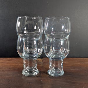 Puede incluir: Dos juegos de copas de cristal transparente, cada uno con una parte superior bulbosa y una base cilíndrica más pequeña. Las copas están apiladas, creando un diseño único de doble capa. El fondo es gris oscuro y las copas están sobre una superficie de madera.