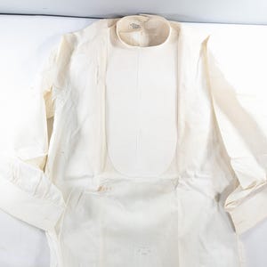 May include: Cream-colored long-sleeved shirt with a rounded collar and a bib-like panel down the front. The shirt has buttoned cuffs and a vertical striped pattern. The fabric appears to be lightweight.