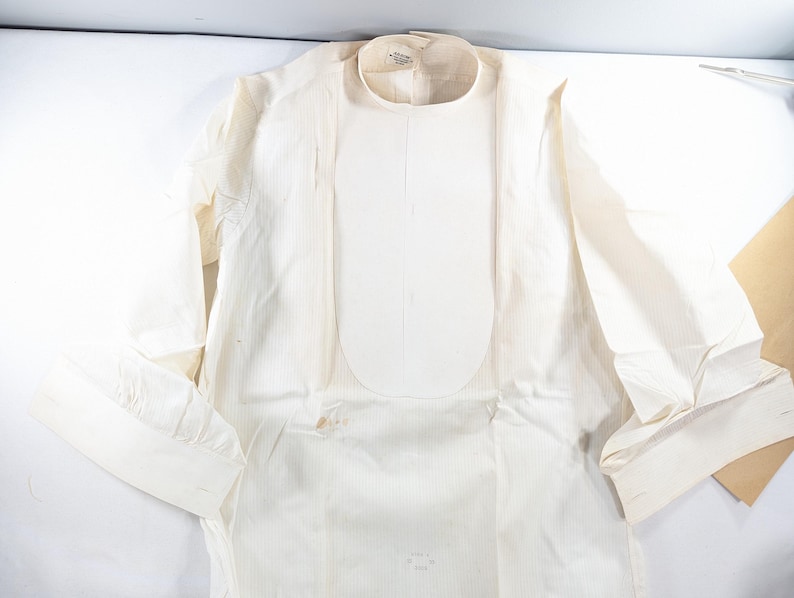May include: Cream-colored long-sleeved shirt with a rounded collar and a bib-like panel down the front. The shirt has buttoned cuffs and a vertical striped pattern. The fabric appears to be lightweight.