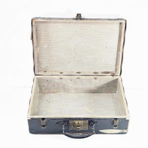 May include: A vintage black suitcase with a rectangular shape, featuring a hinged lid, a central metal latch, and a top handle. The interior is lined with a light-colored, textured material, and the exterior corners are reinforced with metal hardware.