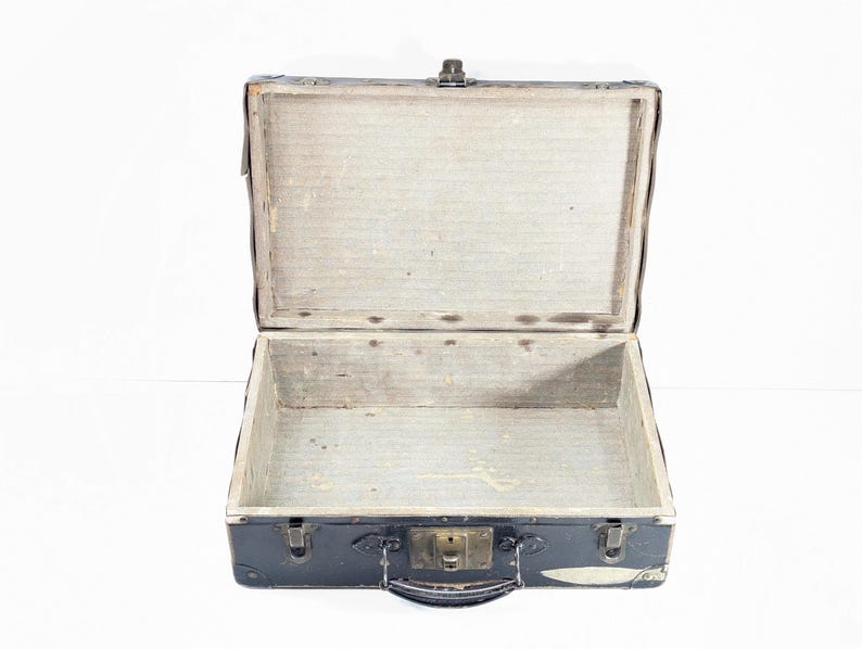 May include: A vintage black suitcase with a rectangular shape, featuring a hinged lid, a central metal latch, and a top handle. The interior is lined with a light-colored, textured material, and the exterior corners are reinforced with metal hardware.