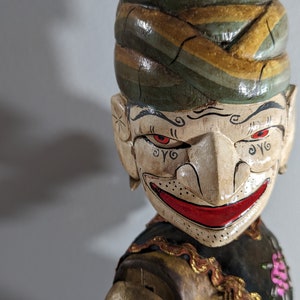Puede incluir: Un títere de madera con la cara pintada, que lleva un turbante de colores. El títere tiene ojos rojos, una amplia sonrisa y detalles intrincados en la cara.