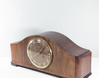 May include: A dark brown wooden mantel clock with a curved top and a rectangular base. The circular clock face features a gold-toned rim, gold-colored Arabic numerals, and gold hands. The brand name Seth Thomas is printed on the clock face.