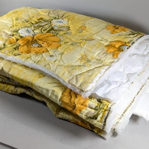 Op de afbeelding: Een gevouwen doorgestikte stof met een geel bloemenpatroon met grote oranje en witte bloemen. Het materiaal heeft een witte tussenvoering zichtbaar aan de randen en bevat tekst langs de zelfkant die luidt Protected with Scotchgard.