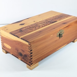 Puede incluir: Una caja rectangular de madera con un diseño floral tallado en la tapa. La caja es de madera marrón clara y oscura, con un cierre de latón. Se apoya sobre cuatro pequeños pies cuadrados.