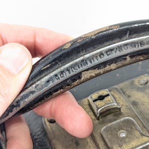 May include: A black carrying handle with visible wear and the embossed text BRITISH MADE. The handle is attached to a dark case featuring a weathered brass-toned metal latch mechanism with a central keyhole and visible mounting rivets.
