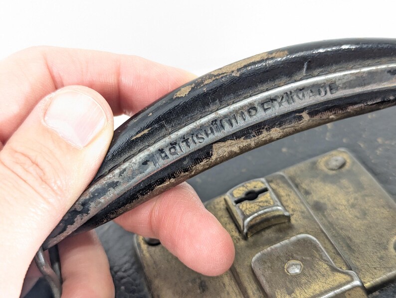 May include: A black carrying handle with visible wear and the embossed text BRITISH MADE. The handle is attached to a dark case featuring a weathered brass-toned metal latch mechanism with a central keyhole and visible mounting rivets.