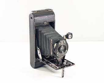 May include: A vintage black folding camera with a bellows mechanism, a circular lens assembly, and a metal drop-down bed. The front plate features the text Eastman Kodak Co. and the word Kodak is printed on a small metal tab extending downward.