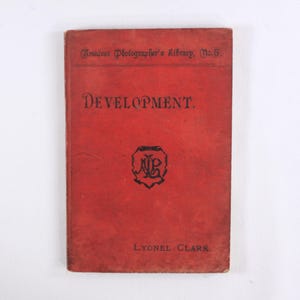 Puede incluir: Libro rojo titulado "Development" con el texto "Amateur Photographer's Library No. 5" y "Lyonel Clark" en la portada.