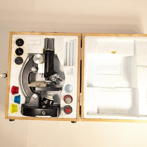Puede incluir: Un microscopio negro con una caja de transporte de madera. El microscopio tiene una base plateada y un brazo negro. La caja tiene un interior de espuma blanca.
