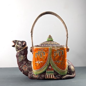 May include: A decorative ceramic camel-shaped teapot. The teapot is primarily brown with orange and green accents. It features a woven handle and is adorned with floral and bird motifs. The teapot measures approximately 10 inches in length.