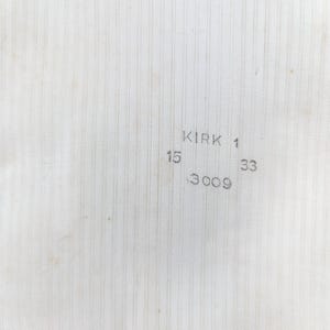 May include: Close-up of a textured, off-white fabric with vertical lines. The text "KIRK 1" is printed in the upper right corner, with the numbers "15", "33", and "3009" below it. The fabric has a slightly aged appearance.