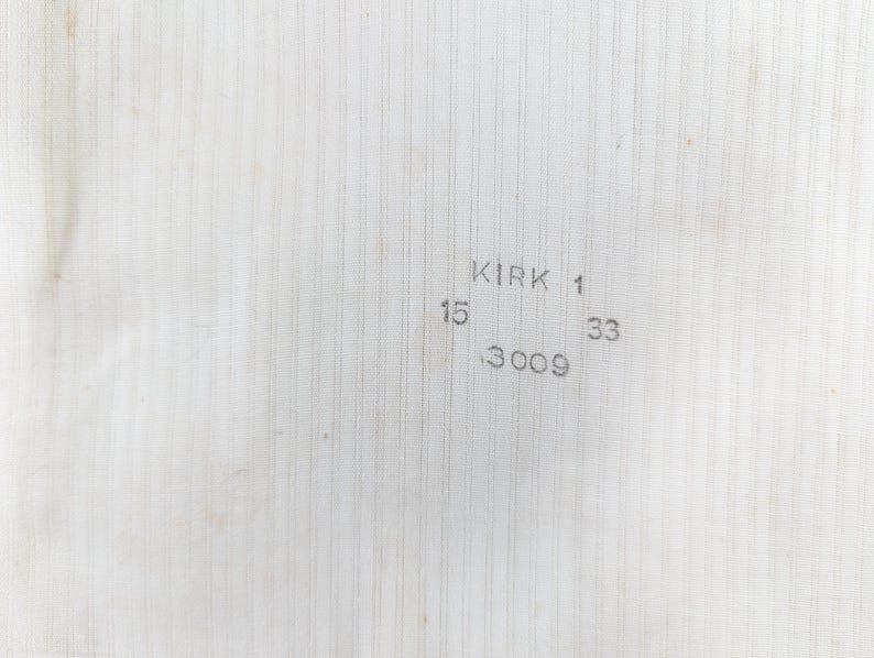 May include: Close-up of a textured, off-white fabric with vertical lines. The text "KIRK 1" is printed in the upper right corner, with the numbers "15", "33", and "3009" below it. The fabric has a slightly aged appearance.