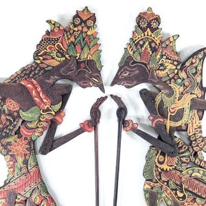 Op de afbeelding: Twee ingewikkeld gesneden Indonesische Wayang Kulit poppen. De poppen zijn bruin met gedetailleerde kleurrijke patronen in rood, groen, geel en goud. Elke pop houdt een houten stok vast en heeft een gedetailleerde hoofdtooi.