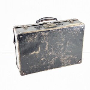 May include: A rectangular black suitcase with a weathered, distressed finish. It features a dark handle, metal corner reinforcements, and a central metal latch mechanism. The surface shows visible signs of wear, including scuffs and scratches.