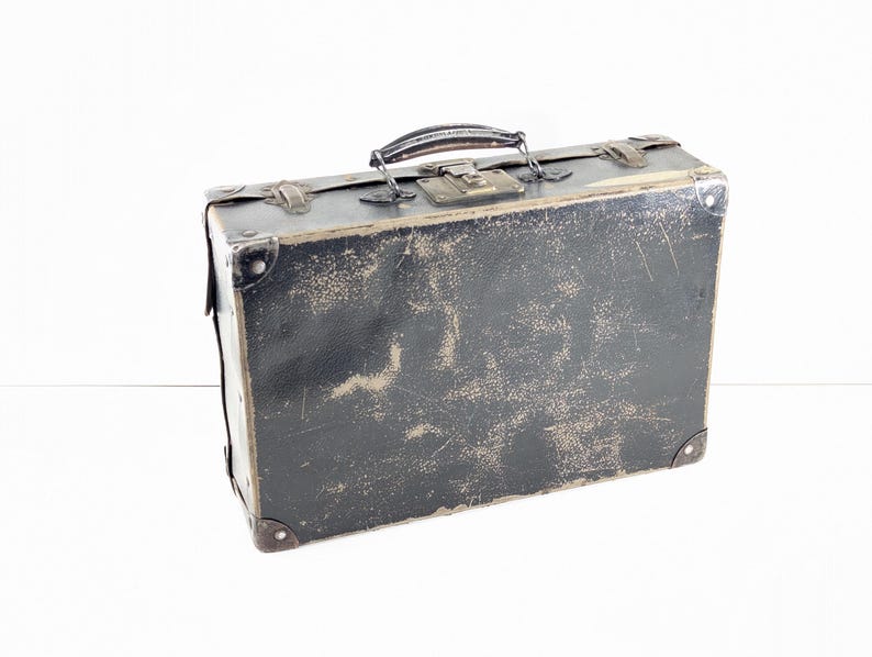 May include: A rectangular black suitcase with a weathered, distressed finish. It features a dark handle, metal corner reinforcements, and a central metal latch mechanism. The surface shows visible signs of wear, including scuffs and scratches.