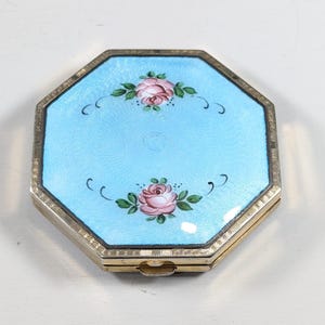 Op de afbeelding: Een achthoekige compacte poederdoos met een lichtblauwe emaille bovenkant. De bovenkant heeft twee roze rozenontwerpen met groene bladeren en zwarte gebogen accenten. De compact heeft een zilverkleurig metalen frame en een sluiting.