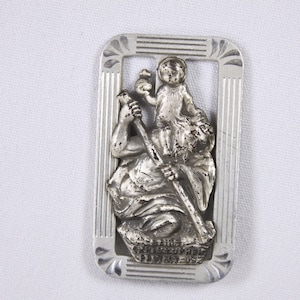 Könnte beinhalten: Silberfarbener Metall-Lesezeichen mit einem Reliefbild des Heiligen Christophorus, der ein Kind trägt. Das Lesezeichen ist rechteckig mit einem dekorativen Rand und der Inschrift "Heiliger Christophorus Beschütze uns" unten.