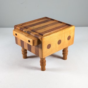 May include: A small wooden cutting board with a striped design and a handle. The board is on four turned legs and has three brown dots on the side.