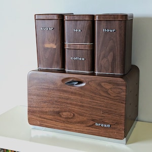 Könnte beinhalten: Ein Vintage-Küchenset mit Holzmaserung. Das Set umfasst einen Brotkasten und Behälter mit den Aufschriften "Zucker", "Tee", "Mehl" und "Kaffee". Der Brotkasten hat einen Griff und das Wort "Brot" ist auf der Vorderseite aufgedruckt.