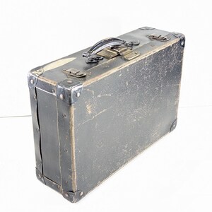 May include: A rectangular, dark gray vintage suitcase featuring a textured exterior with visible signs of wear. It includes a top handle, metal corner reinforcements, and two metallic latches on the upper edge for secure closure of the case.