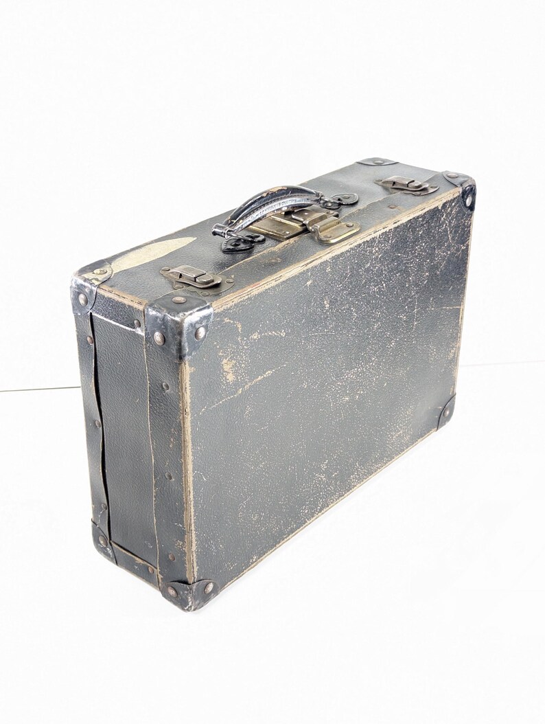 May include: A rectangular, dark gray vintage suitcase featuring a textured exterior with visible signs of wear. It includes a top handle, metal corner reinforcements, and two metallic latches on the upper edge for secure closure of the case.
