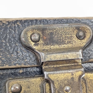 May include: A weathered black textured surface with a tarnished brass metal latch. The latch features embossed text that reads GB PAT 427897 AND OTHERS MADE IN ENGLAND, secured by two rivets. The metal shows signs of age, oxidation, and wear.