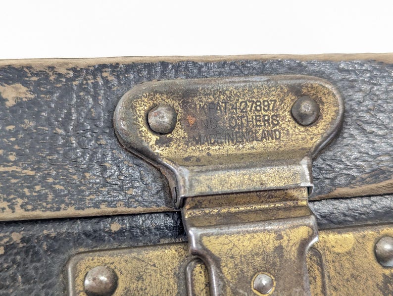 May include: A weathered black textured surface with a tarnished brass metal latch. The latch features embossed text that reads GB PAT 427897 AND OTHERS MADE IN ENGLAND, secured by two rivets. The metal shows signs of age, oxidation, and wear.