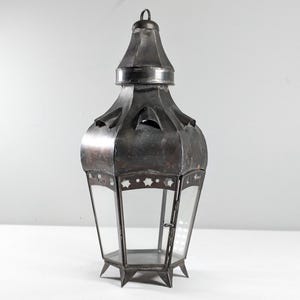 May include: A dark metal lantern with a decorative design. The lantern has a hexagonal shape with clear glass panels and a hinged door. The top features a loop for hanging. The lantern is suitable for candles or lights.