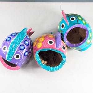 Peut inclure: Trois nichoirs en céramique en forme de poisson peints à la main. Les nichoirs sont peints dans des tons de bleu, de violet, de rose et de jaune, avec des motifs décoratifs. Chaque nichoir a une grande ouverture et un petit détail d'aileron.