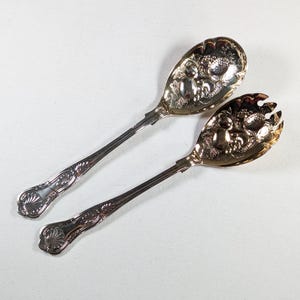 May include: A pair of silver-toned metal salad serving utensils consisting of one solid spoon and one slotted spoon. Both utensils feature ornate embossed fruit and leaf designs on the bowls and decorative scrollwork on the handles.