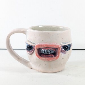 Puede incluir: Una taza de cerámica con esmalte blanquecino moteado que presenta un rostro esculpido en la parte frontal. El rostro tiene labios rosados prominentes, ojos oscuros hundidos y dientes pequeños, oscuros y texturizados en la boca abierta.