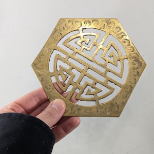 May include: A hexagonal brass trivet featuring an intricate openwork design in the center and a decorative etched border pattern along the edges. The metallic surface has a polished gold-toned finish with visible engraved details throughout the piece.