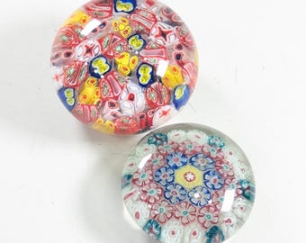 May include: Two round glass paperweights featuring intricate millefiori patterns. The top paperweight displays a dense arrangement of red, yellow, and blue floral-like glass canes. The bottom paperweight shows a symmetrical, geometric floral design in shades of pink, blue, yellow, and white encased in clear glass.
