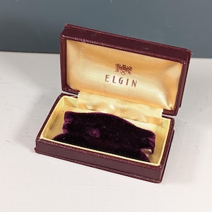 May include: Open burgundy jewelry box with a gold satin interior and a dark purple velvet insert. The box is rectangular with a hinged lid. The word "ELGIN" is printed in gold on the inside of the lid.