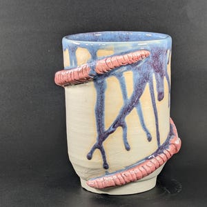 Op de afbeelding: Een handgemaakte keramische beker met een lichtbeige basis, blauwe rand en paarse druppels. De beker heeft een getextureerd, roze, wormachtig ontwerp dat om de buitenkant gewikkeld is. De binnenkant is lichtblauw.
