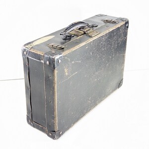May include: A rectangular black suitcase with a weathered, textured surface. It features a dark handle, metallic corner reinforcements, and two metal latches on the top edge. The exterior shows signs of wear with visible scuffs and scratches.