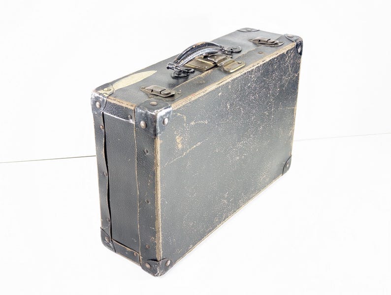 May include: A rectangular black suitcase with a weathered, textured surface. It features a dark handle, metallic corner reinforcements, and two metal latches on the top edge. The exterior shows signs of wear with visible scuffs and scratches.