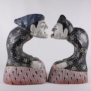 Peut inclure: Une paire de figurines en bois représentant un homme et une femme, tous deux vêtus de vêtements traditionnels et agenouillés, la tête baissée. Les figurines sont peintes en noir, blanc, rose et bleu.