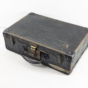 May include: A rectangular black suitcase with a weathered surface, featuring reinforced metal corners, a central brass-toned latch, and two side locking mechanisms. The case includes a black handle attached to the front with metal hardware.
